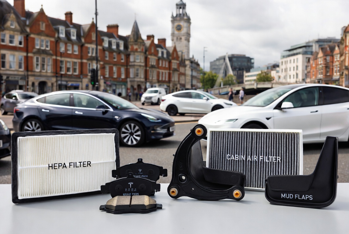 Assortment of essential Tesla parts including HEPA filter, brake pads, control arm, cabin air filter, and mud flaps, displayed on a table with Tesla vehicles and a UK cityscape in the background